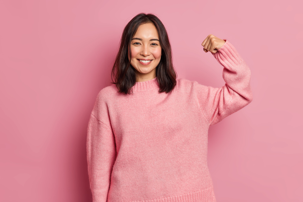 powerful cheerful brunette woman raises arm shows muscle demonstrates her strength looks confident camera smiles gently wears casual long sleeved jumper